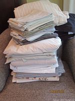 A stack of assorted bedding items including pillow cases and bed sheets with various colors and patterns piled on an armchair.