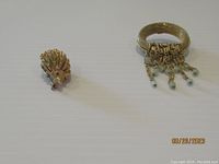 Two brooches on a white surface, one is a porcupine with pink eyes, the other is a round gold-tone piece with dangling beads.