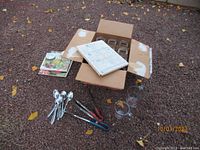Top view of open box showing 12 Ball Mason jars inside, two books, four spoons, two pairs of tongs, and two glass measuring cups arranged outside the box on gravel ground.