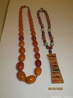 Two large beaded necklaces on a fabric surface, one with large orange and maroon beads and the other with alternating frosted spherical beads and a rectangular pendant.