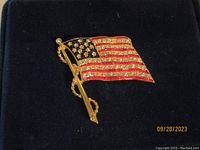 Close-up image of the American flag brooch showing gold-tone metal with rhinestones and red enamel stripes, on a dark blue background.