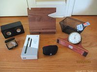Overview of all lot items on wooden floor: two clocks, Neo Smart Pen box, Bluetooth speaker, wire basket, wooden cell phone holder, small black pouch likely for Logitech QuickCam.