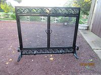 Full front view of the Plow & Hearth metal fireplace screen with scrollwork design, showing the two doors and mesh panels on a gravel background.