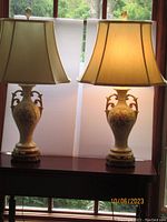 Pair of vintage porcelain table lamps with beige fabric bell shades. Positioned indoors against window.