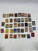 Top down view of 40 vintage assorted collectible advertising matchbook covers and match boxes arranged in rows showing variety of brands and colorful designs.