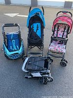 Four assorted baby strollers placed on outdoor pavement including a blue stroller with canopy, a pink and gray geometric pattern stroller with tray, a compact black and gray stroller folded, and a blue stroller with a seating area.