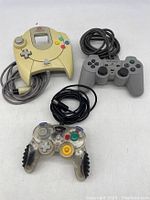 Three game controllers shown together: Sega Dreamcast, Sony PlayStation SCPH-1200, and MadCatz translucent GameCube controller on white background.