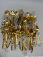 Full view of the gold plated flatware set laid out showing multiple forks, spoons, knives, and serving utensils with bamboo style handles, some still in plastic packaging.
