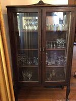Front view of wooden glass display cabinet showing two glass doors, metal handles, and interior shelves with glassware (not included).