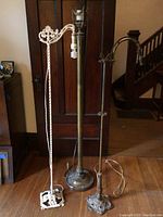 Two vintage metal floor lamps side by side in front of a wooden door. One lamp is dark metal with simple circular base, the other is white with an ornate chain and decorative base. Both show wear and wires are visible.