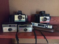Front view of five vintage cameras including Polaroid Swingers, Polaroid Colorpack II, and Kodak Instamatic 14 models on shelf.