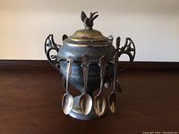 Front view of metal urn showing four attached spoons and decorative lid with bird figurine. Surface shows scratches and discoloration.