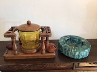 Photo showing a wooden pipe stand with yellow glass container and wooden lid, two wooden pipes resting on the stand, and a blue-green carved stone ashtray beside it on a wooden surface.