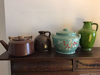 Image showing four vessels on a wooden surface against a plain wall: a brown metal teapot with wooden handle, a dark brown ceramic jug, a blue ceramic pot with floral design and lid, and a green ceramic pitcher.