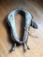Top-down photo of a used antique horse collar saddle with worn leather and metal ball attachments, placed on a wooden floor.