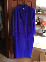Full front view of a deep purple vintage women's long double-breasted coat hanging on hanger against a wooden door. Shows coat length, button placement, and pocket design.