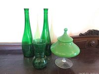 Full view of four green glassware items showing two tall bottles, one textured vase, and one covered candy dish on a pedestal.