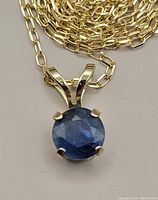 Close-up of the round sapphire pendant set in yellow gold with four prongs, showing gemstone color and facets.