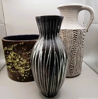 Photo showing the three vases together from the front: the largest tall white textured vase with handle in the back right, a middle-sized dark vase with vertical white stripes in front center, and a shorter brown planter with lime-green glaze drips on the left.
