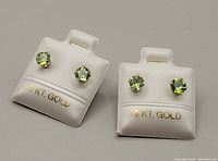 Close-up of two pairs of 10kt white gold stud earrings with light green peridot stones, one larger pair and one smaller pair, displayed on white holders marked 10kt gold