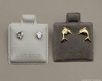 Two pairs of screw-back earrings displayed on grey and white velvet earring holders, one white holder with heart-shaped cubic zirconia earrings in white gold, the other grey holder with yellow gold dolphin-shaped earrings.