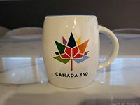 White Canada 150 commemorative coffee mug with multicolored maple leaf logo and text 'CANADA 150' showing front design and handle.