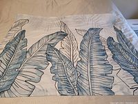 Full view of the cotton wall tapestry laid out flat on a bed showing blue and gray stylized banana leaves on a white background.
