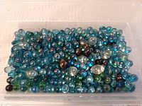 Clear plastic container filled with assorted decorative glass beads in shades of blue, green, and iridescent tones.