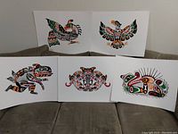Photo showing five John Nelson First Nations art prints laid out on a couch, including art depicting a Sea Monster, Otter and Sea Urchin, Longhouse Mask, Thunderbird, and Eagle in traditional Indigenous style with vibrant colors.
