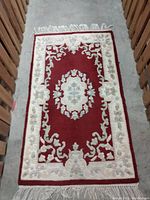 Full view of red and cream rectangular rug showing floral medallion and border pattern, placed on grey carpet.