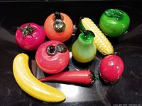Nine colorful glass fruit and vegetable figurines displayed on black surface. Items include banana, apple, tomato, corn, carrot, pear, and others.