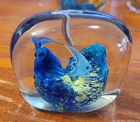 Clear glass paperweight with a blue coral formation and a small fish inside, shown from the front on a wooden surface.