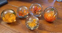 Photo showing five spherical glass paperweights with unique orange floral designs inside on a wooden surface.