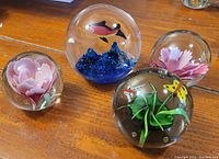 Four glass paperweights on wooden surface: one large with underwater sea scene, two smaller clear glass with pink flowers, and one spherical with flowers and butterflies