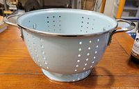 Side view of pale teal metal colander showing perforations, two handles, and round base on a wooden table