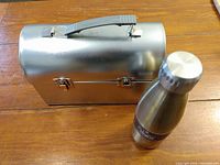 Photo of silver metal lunch box with handle and latch closures beside silver stainless steel thermos.