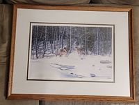Framed wolf print showing two wolves in snowy woodland, natural wood frame, white matte visible.