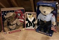 Three teddy bears in original packaging displayed together; a military-themed brown bear, a regal small bear, and a white uniformed bear