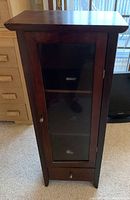 Front view of the tall wooden cabinet with a glass door closed, showing the dark wood finish and the drawer at the bottom.