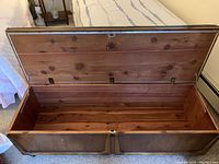 Open cedar chest showing smooth, reddish natural cedar wood interior with Lane brand mark on underside of lid. Lock on front. Chest sits on wheels and has black cushioned top.