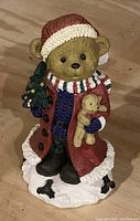 Ceramic bear figurine dressed in winter clothing holding a Christmas tree and a smaller bear toy, detailed with a red coat and scarf