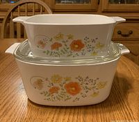 Two Corning Ware casserole dishes stacked; one has orange and yellow flowers, other has no lid but same pattern as bottom dish