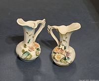 Two small ceramic pitchers with floral relief detail, handle and scalloped rims, pastel colors.