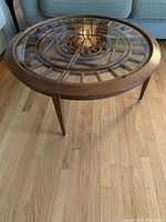 Angle view of round coffee table showing wooden frame, tapered legs, and glass top with metal design underneath.