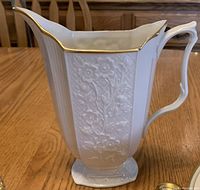 Porcelain pitcher with embossed floral pattern and gold trim, side view showing elegant handle.