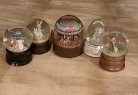 Seven musical globes arranged in a row on a wooden surface, showing different figurines inside clear glass spheres and domes. Varied shapes including round and teardrop glass covers. Bases mostly dark wood or brown. Figurines include rabbits, children, carousel, and animal riders.