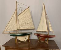 Two wooden model sailboats on stand, one with green hull and one with red hull, off-white canvas sails