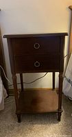 Photo showing dark wood nightstand with two drawers, round ring pull handles, tapered legs, and a lower shelf placed on carpeted floor, positioned between bed and wall.