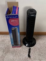 Photo of the black oscillating tower fan beside its original open box showing the fan's front and side profile.