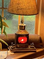 Lit lamp showing metal vintage ship lantern style base with red light glowing behind glass, topped with burlap lamp shade, on nautical rope trimmed wooden base.